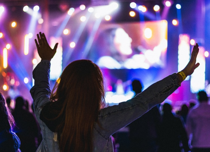 A person with their hands in the air watching a large screen in a crowded faith auditorium.