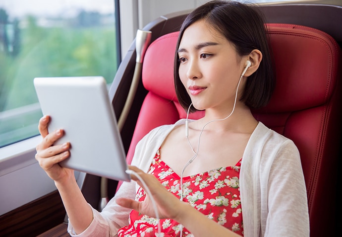 An Asian female sitting on a train, smiling and looking at her pad