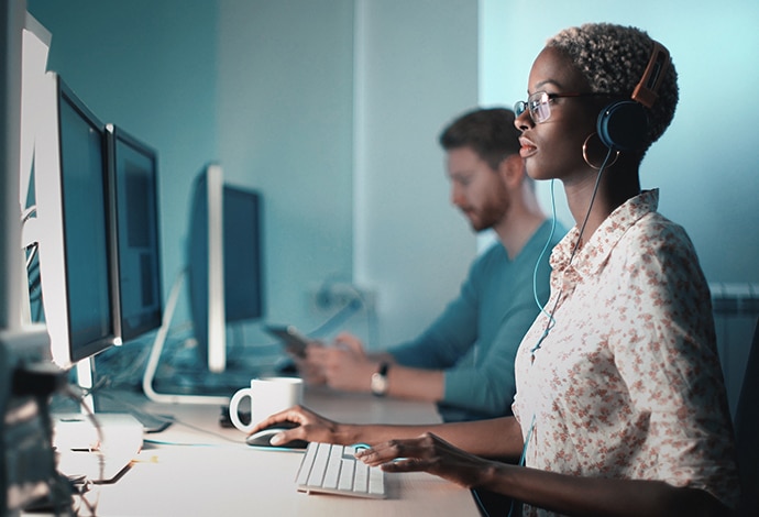 A female sitting at a white desk, looking at her monitor and typing on a keyboard with her headphones on