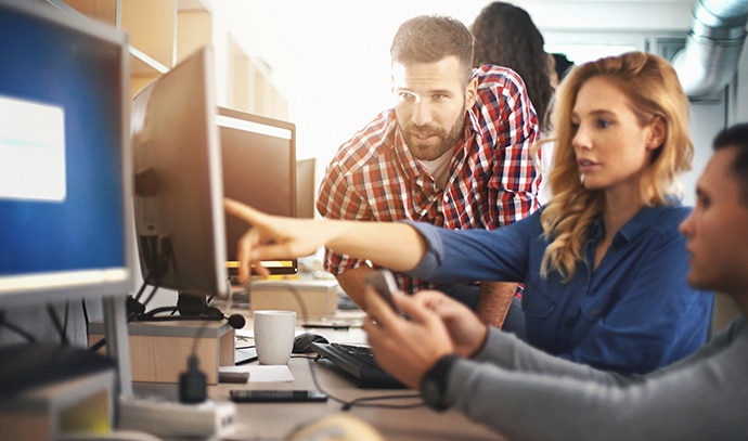 A blond female at her desk with a male on either side, pointing at the monitor screen and talking to the males