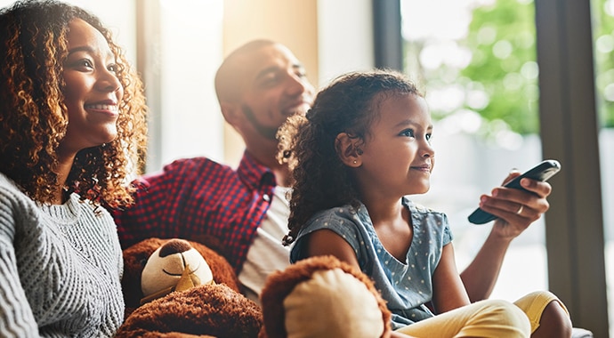 A female with curly hair and her partner on a sofa, with their daughter in between, sitting on a sofa with a larger teddy bear. The male is holding a remote control. They all seem to be smiling in the direction of the TV