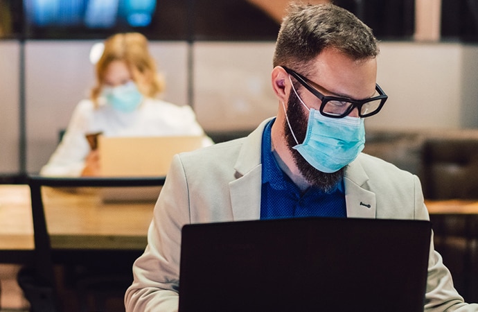 Man sat at a desk working on a laptop. He is wearing a facemask. Behind him is a woman, sat at a safe distance, also wearing a mask.