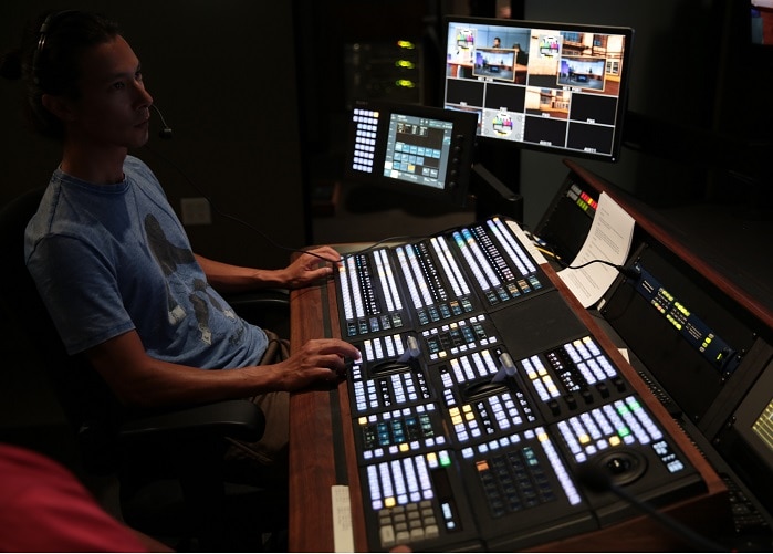 Man operates AV mixing desk in KLCS Control Room. He is seated at extreme left of image, with the desk and controls in front of him. View #3.