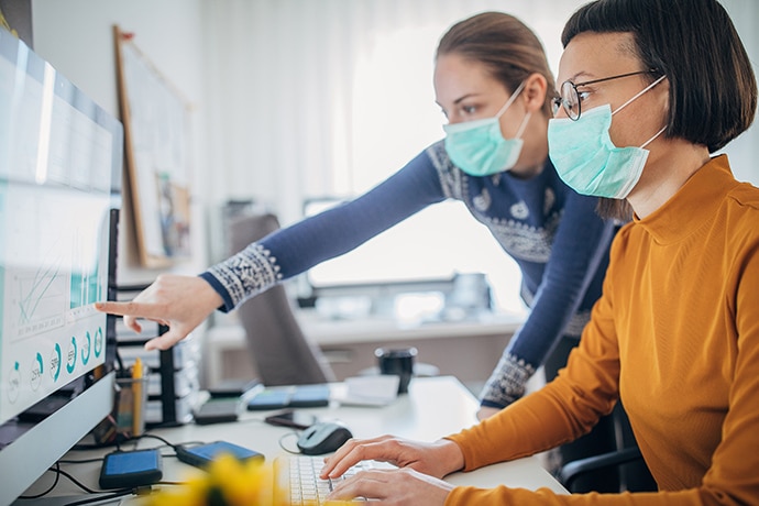 Two women working together at a computer monitor while wearing masks.