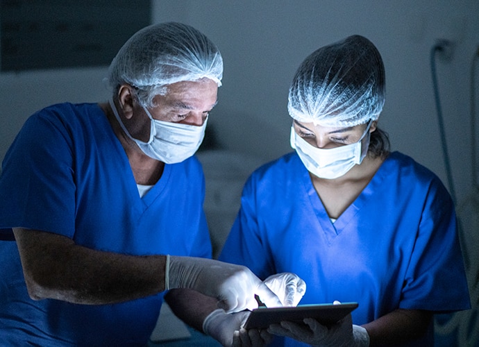 Two masked surgeons pointing at a tablet screen while speaking