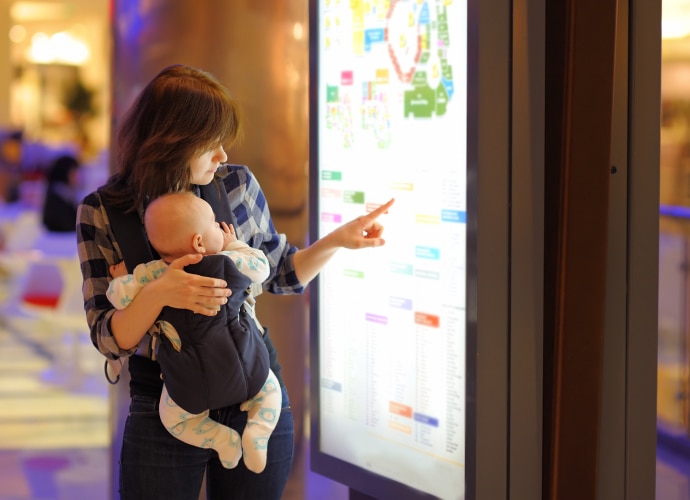 a woman with a baby touches screen to find her way around a shopping mall