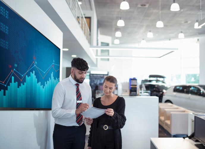 Two staff members in car dealership talk with a display on the wall with a graphic