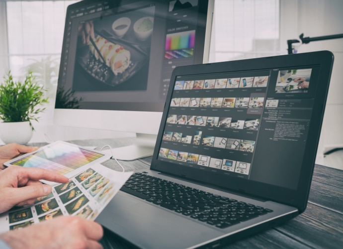 Hands holding a paper with images and laptop and monitor in the background.