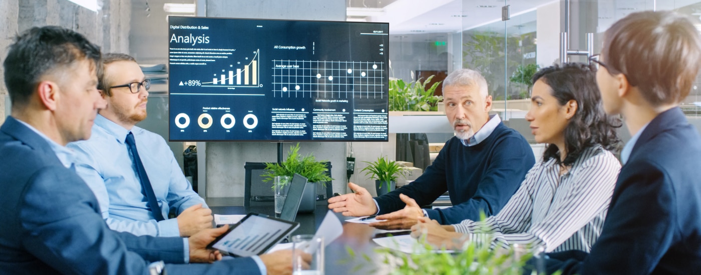 Office meeting room with 5 people talking and large display showing business information