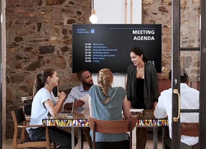 Konferenzraum mit Teilnehmenden in einer Gesprächsrunde und einer Tagesordnung auf einem großen Display-Bildschirm