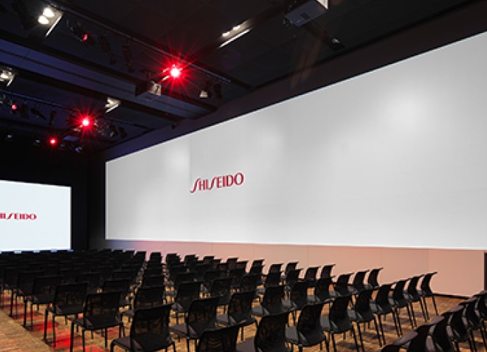 Un display Crystal LED è posizionato nella parte anteriore di una sala buia. Ci sono diverse file di sedie e il logo Shiseido su un ampio schermo di proiezione sul lato della sala.